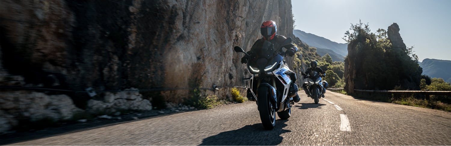 Two motorcyclists riding on a winding mountain road with rocky cliffs and trees under a clear sky
