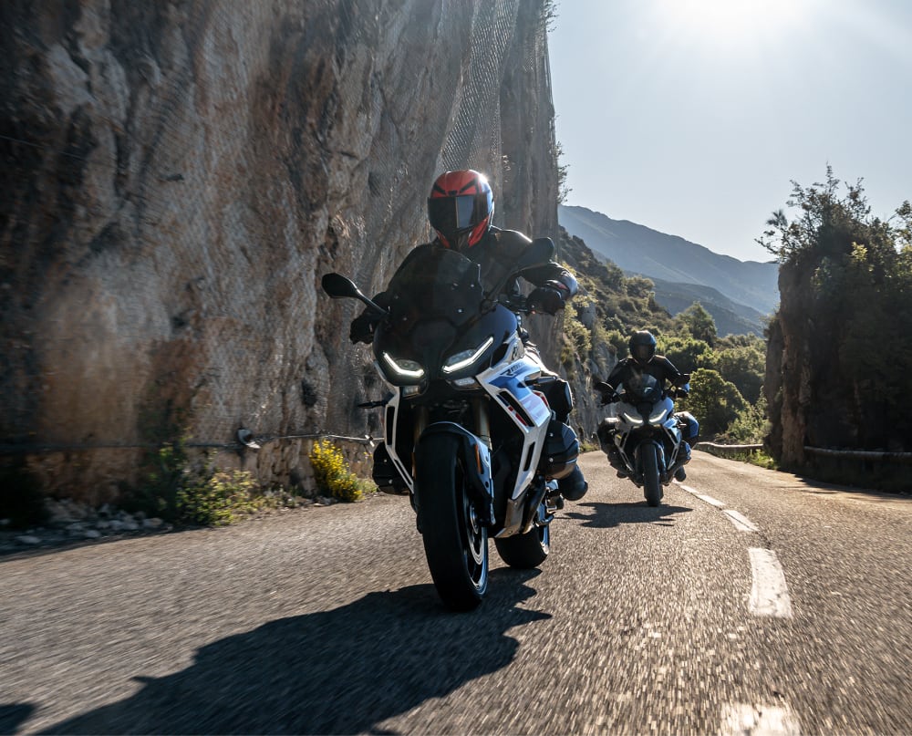 Two motorcyclists riding on a winding mountain road with rocky cliffs and trees under a clear sky