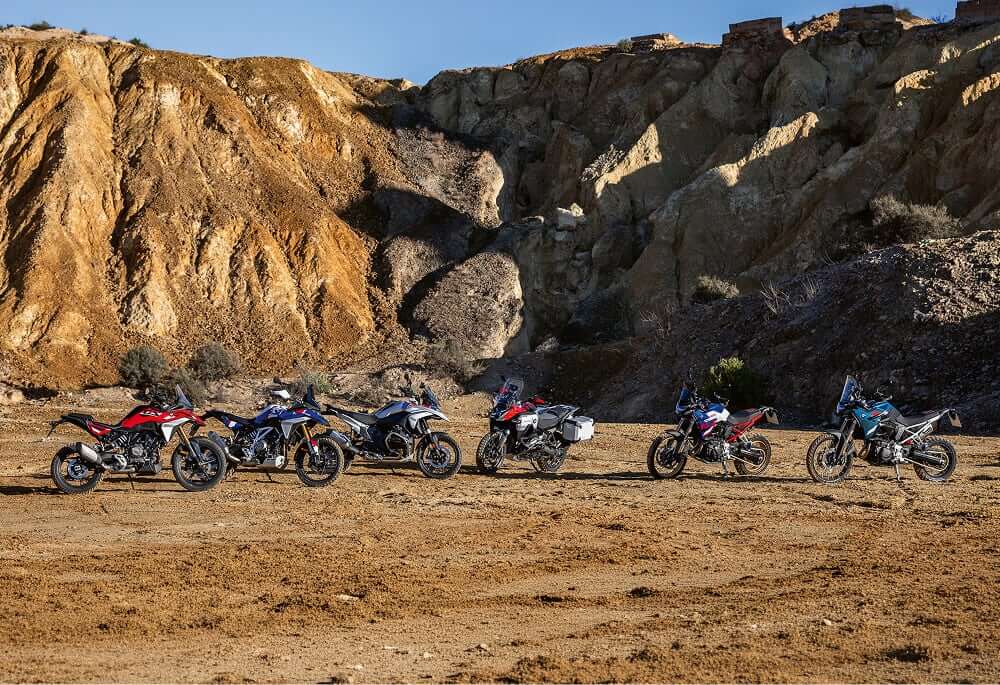 Six motorcycles parked side by side on a dry, rocky terrain with steep, sunlit cliffs in the background