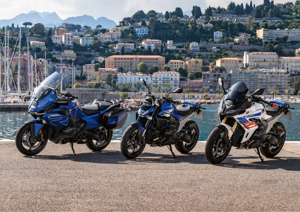 Three BMW motorcycles parked by a waterfront with a marina and hillside town in the background on a sunny day