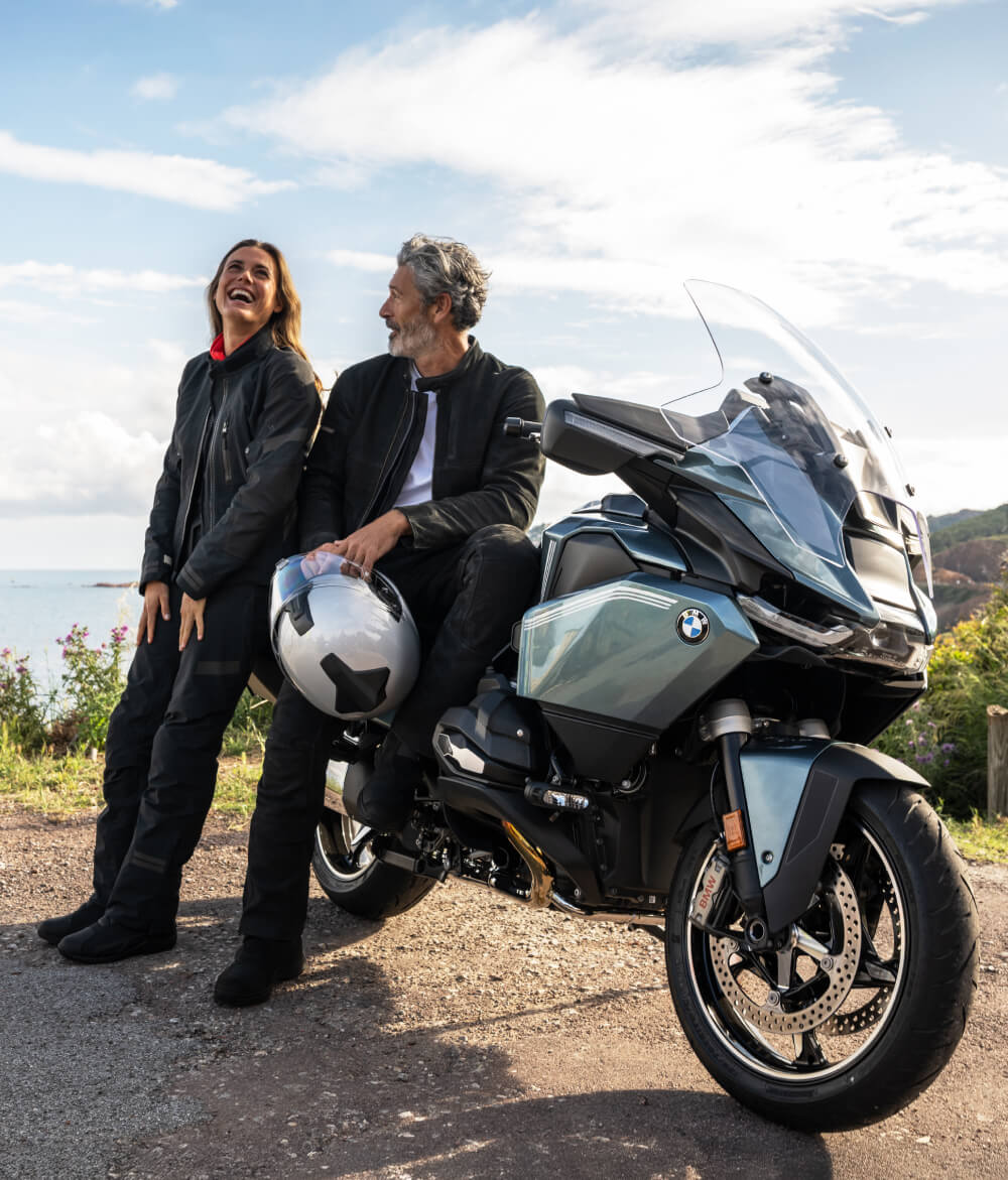 Two people in motorcycle gear smiling by a BMW motorcycle on a sunny, scenic roadside