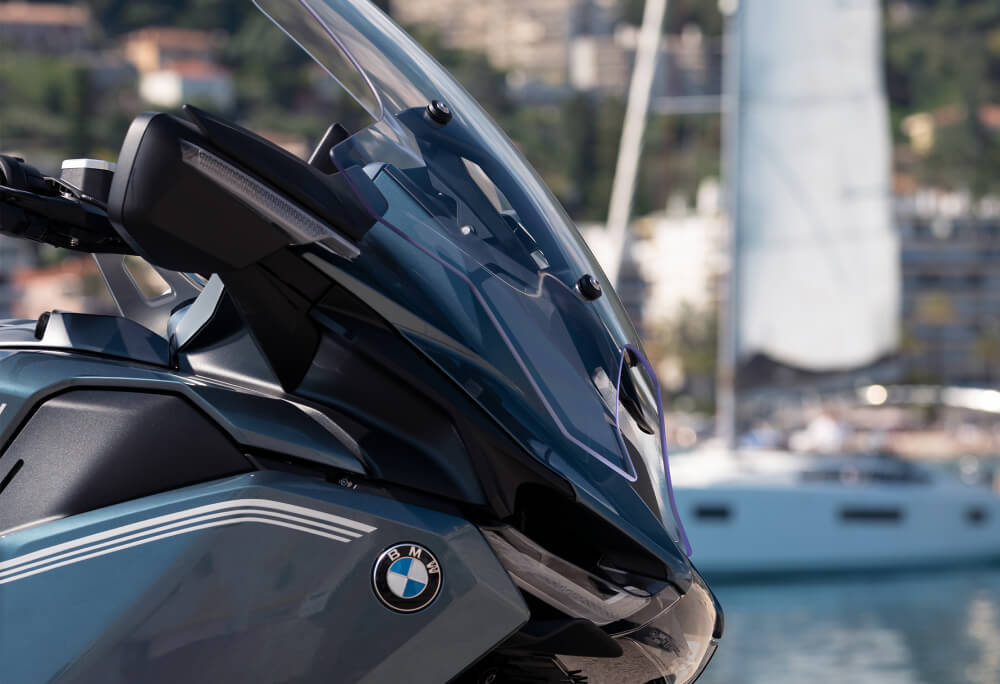 Close-up of a blue BMW motorcycle front with clear windshield, parked near a docked sailboat on a sunny day