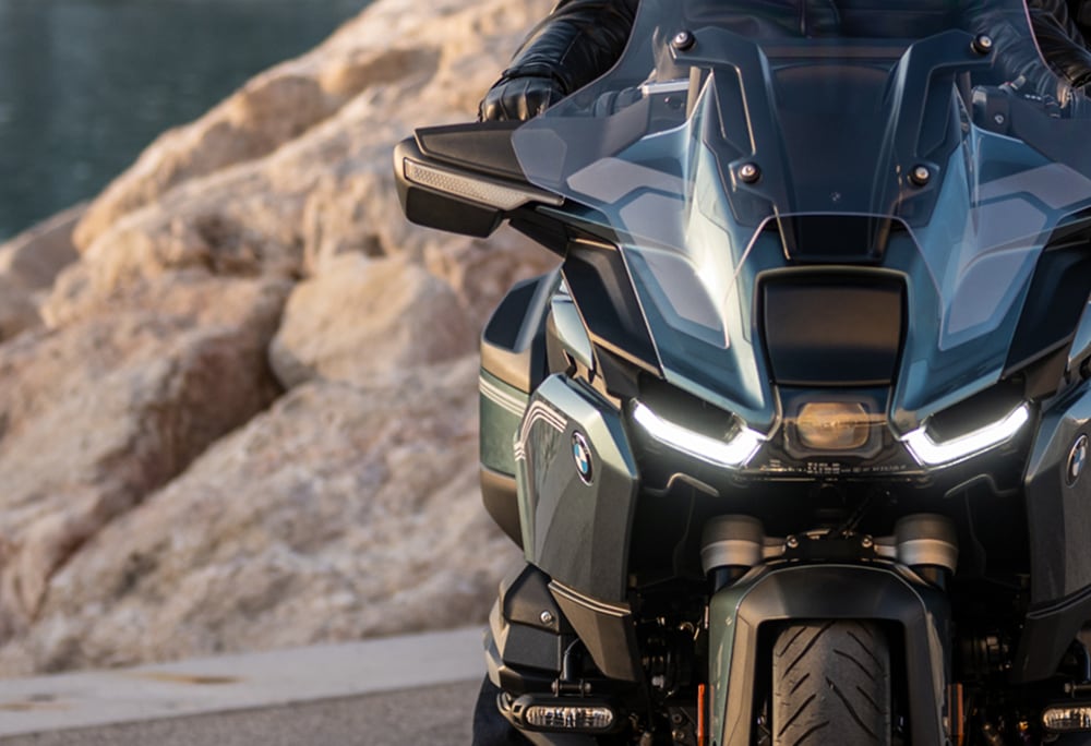 Front view of a BMW motorcycle with illuminated headlights and a tinted windshield, parked near rocky terrain