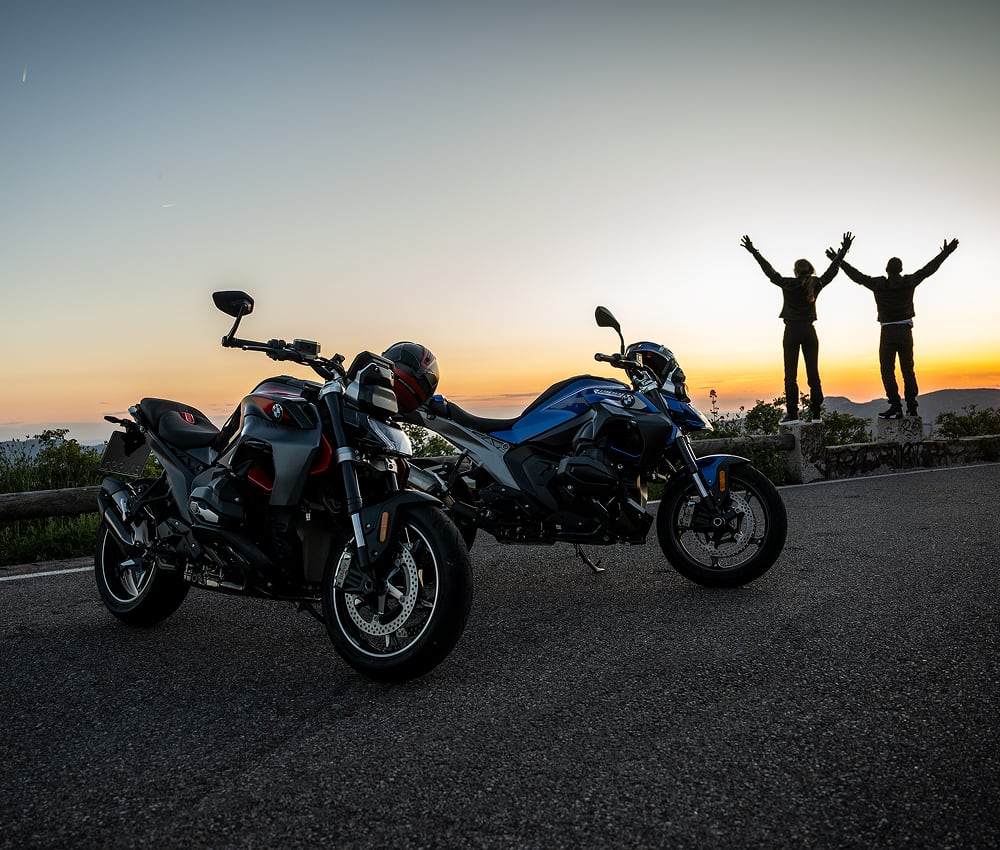 Front side angled view of two BMW motorcycles with two people standing with hands in the air