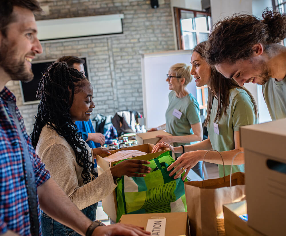 A diverse group of smiling volunteers sorting donations in cardboard boxes at an indoor community event, with name badges visible and donated items in the background.