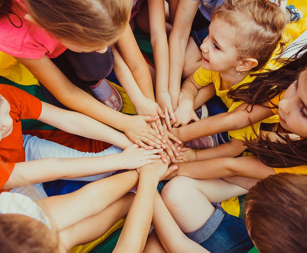 A group of young children sitting in a circle outdoors, reaching their hands into the centre in a team huddle gesture, symbolising teamwork and togetherness.