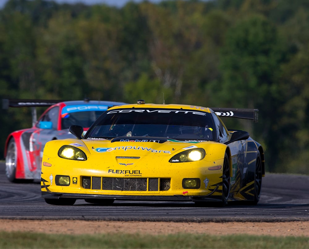 Yellow Corvette race car leads a red race car on a curved track with a blurred forest background