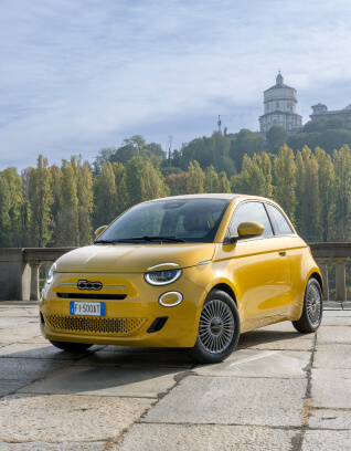Yellow Fiat 500 Hybrid parked on a stone pavement with tall trees and a historic tower in the background under a partly cloudy sky