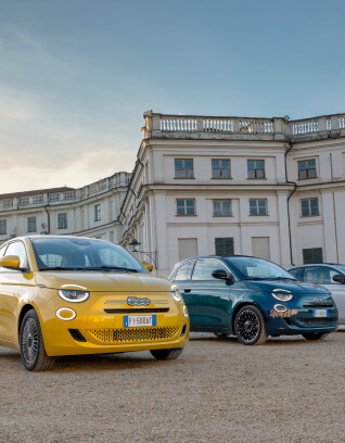 Two Fiat 500 Hybrid cars, one yellow and one dark green, parked on a gravel area in front of a large historic white building under a clear sky