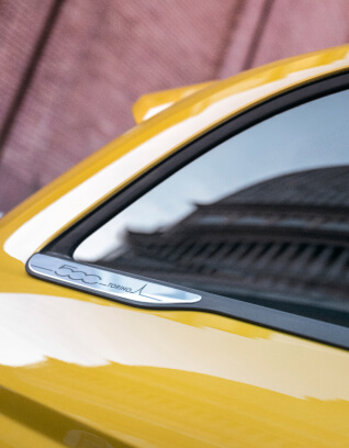Close-up of a yellow Fiat 500 Hybrid badge near the rear side window reflecting a building