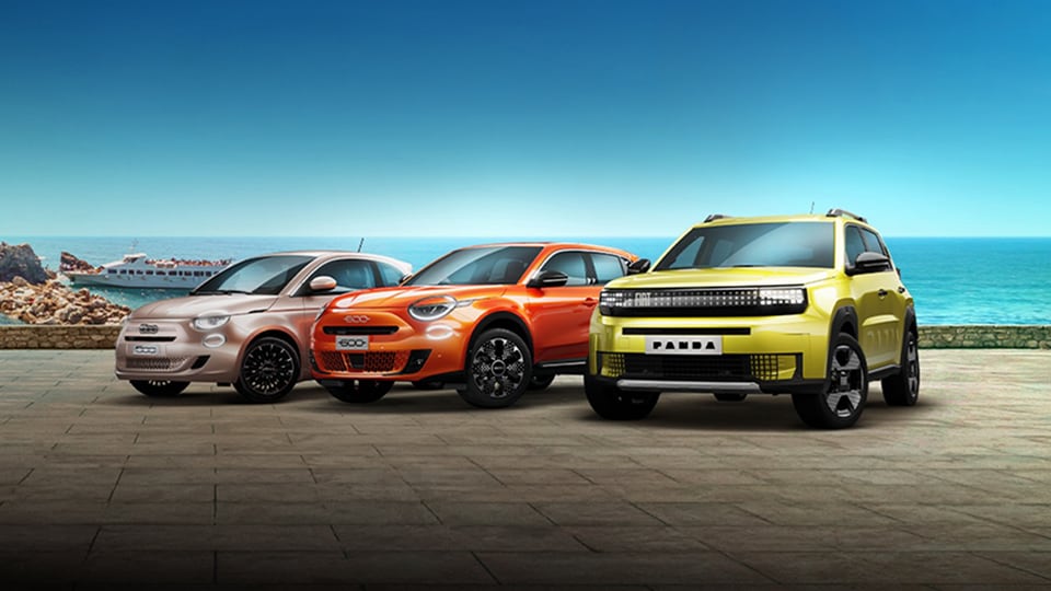 Three colorful Fiat cars parked on a seaside promenade, with a rocky cliff, boat, and bright blue ocean in the background.