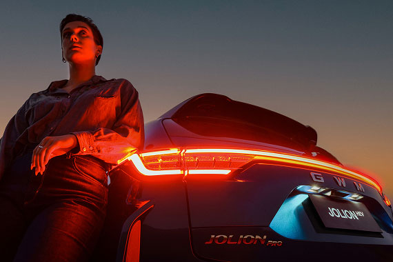 Woman in a denim jacket leans against the Haval Jolion with glowing red taillights at dusk