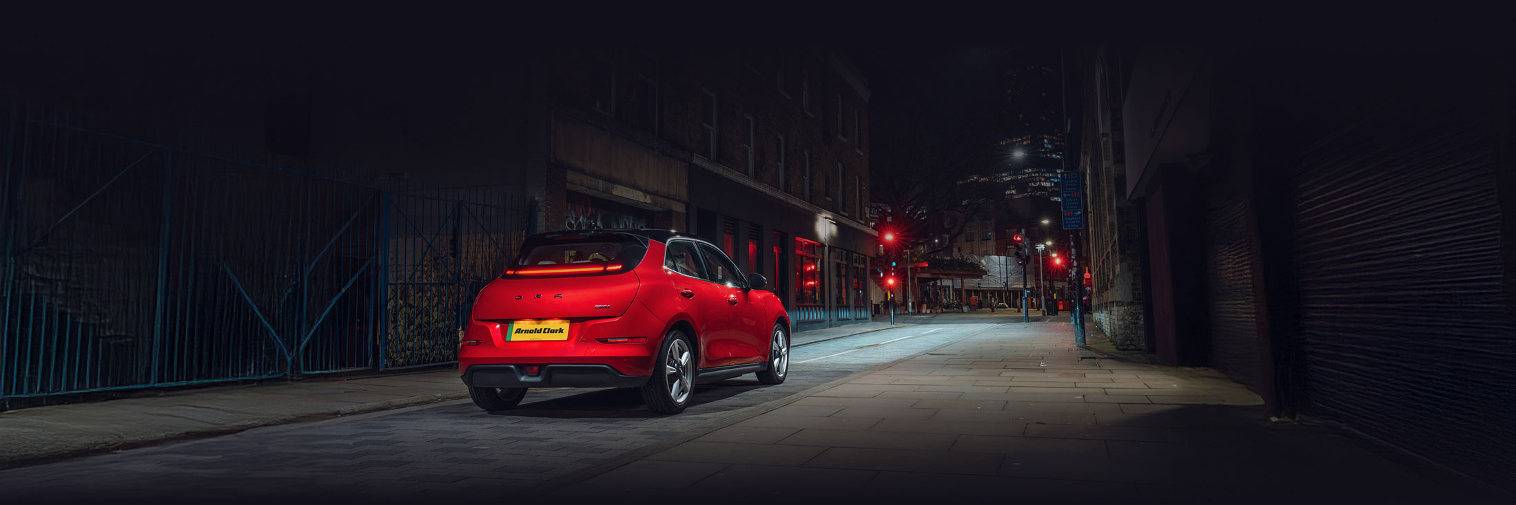 Rear view of a red GWM ORA 03 on a city street at night with headlights on