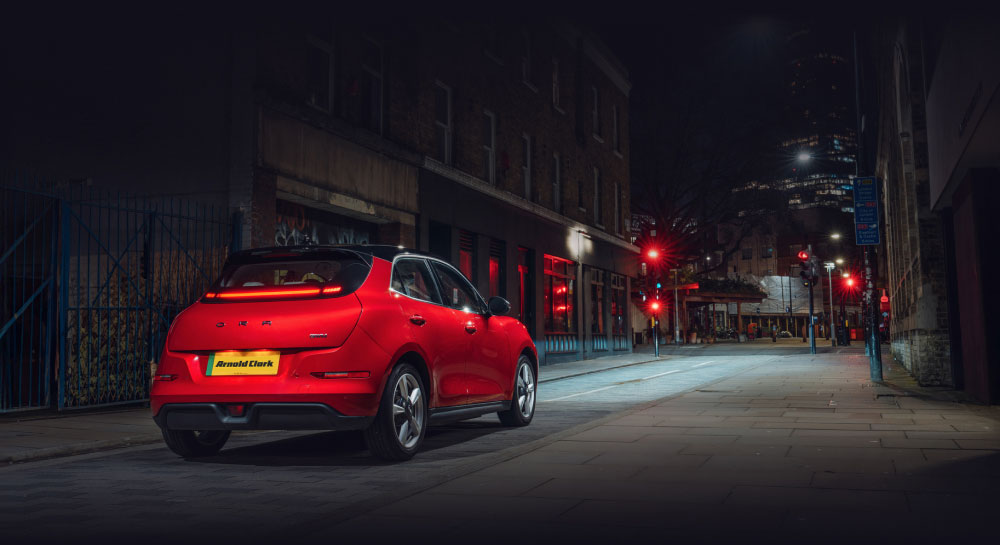 Rear view of a red GWM ORA 03 on a city street at night with headlights on