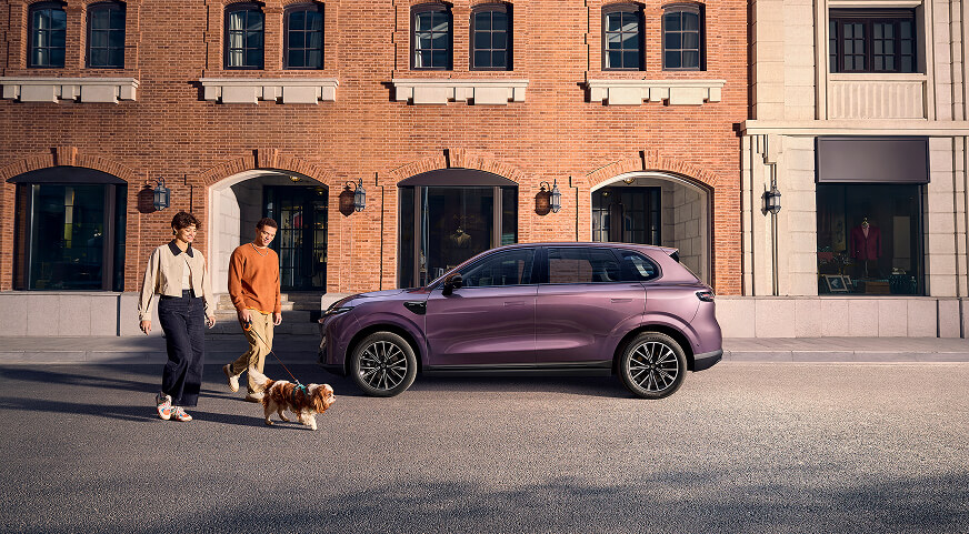 Two people walk a dog past a parked purple Leapmotor B10 in front of a brick building with arched windows and doors
