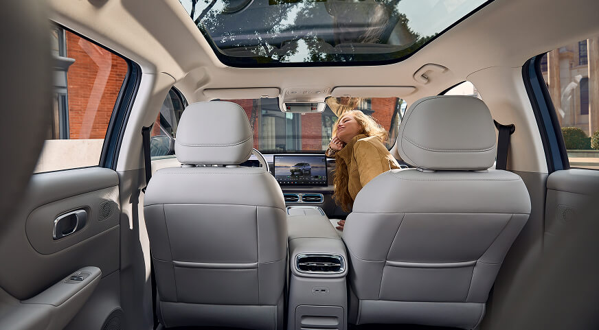 View from the back seat showing light gray leather seats, a panoramic sunroof, and a woman leaning inside the front passenger side of a Leapmotor B10