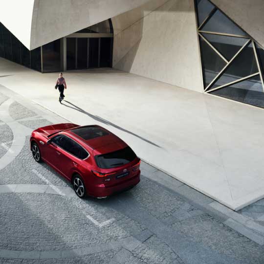 Image of woman in a city walking past a red Mazda CX-60 and casting a long shadow on the pavement
