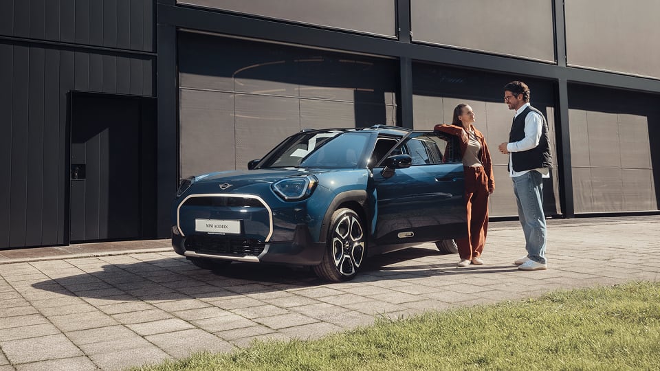Two people stand beside a blue Mini Aceman parked on a paved driveway near a modern building