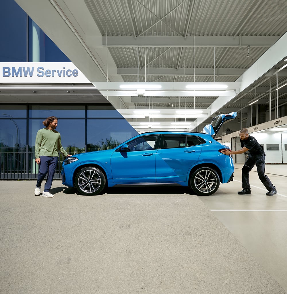 Two men with a blue BMW SUV at a BMW Service center, one standing outside and one opening the trunk indoors
