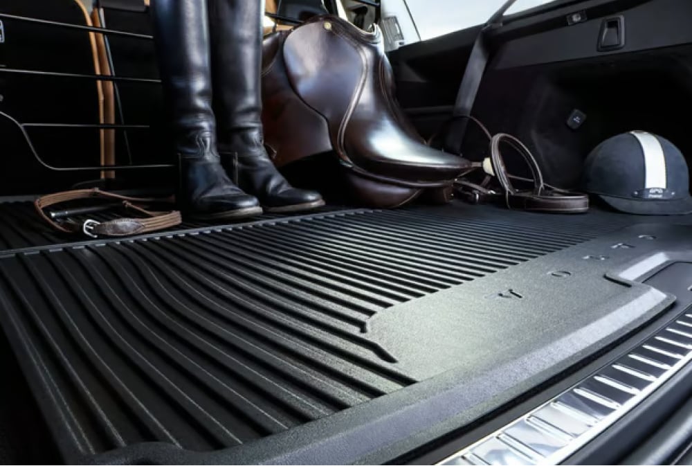 Car trunk with black rubber mat, showing equestrian gear including tall riding boots, saddle, reins, and riding helmet