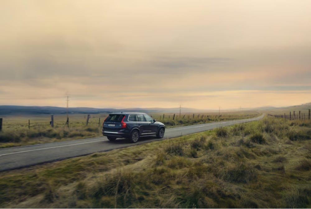 Black SUV driving on a winding country road through grassy fields under a cloudy sky at sunset