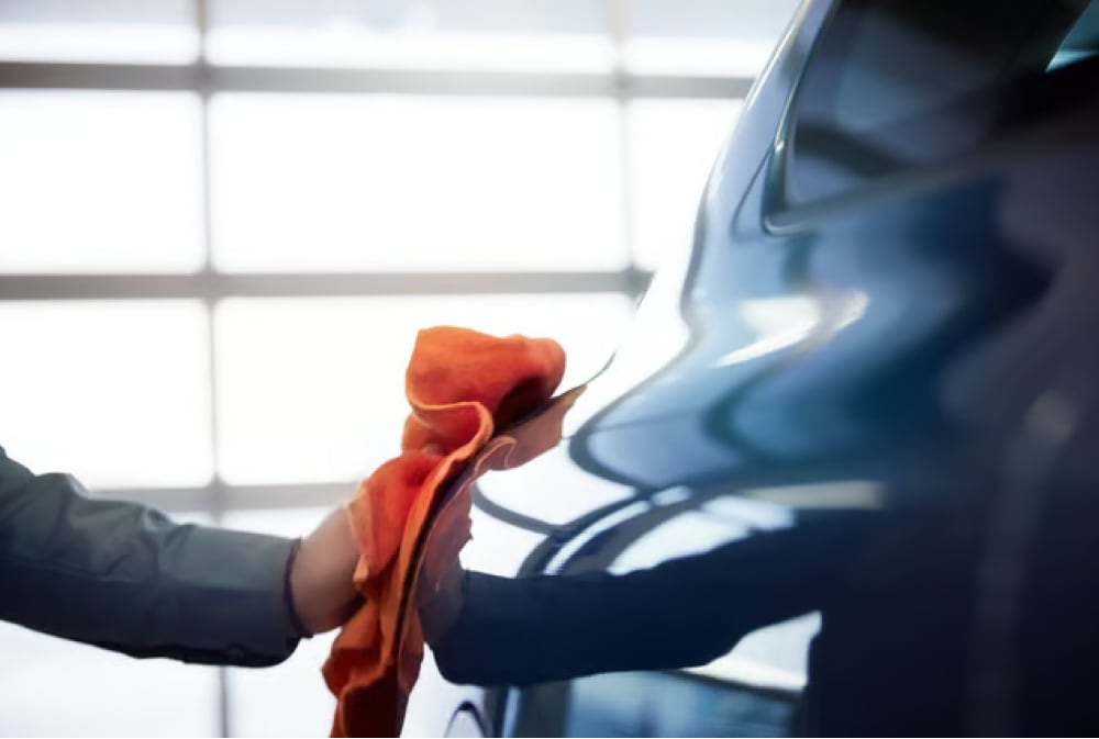 Person cleaning a shiny blue car with an orange cloth in a brightly lit garage