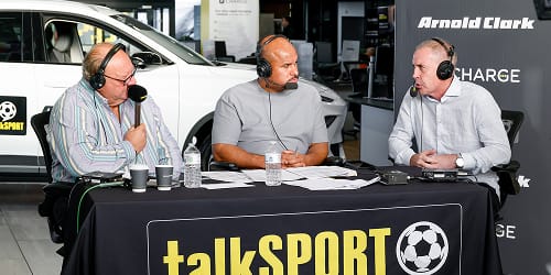 Three people wearing headsets at a talkSPORT broadcast table in a car showroom.