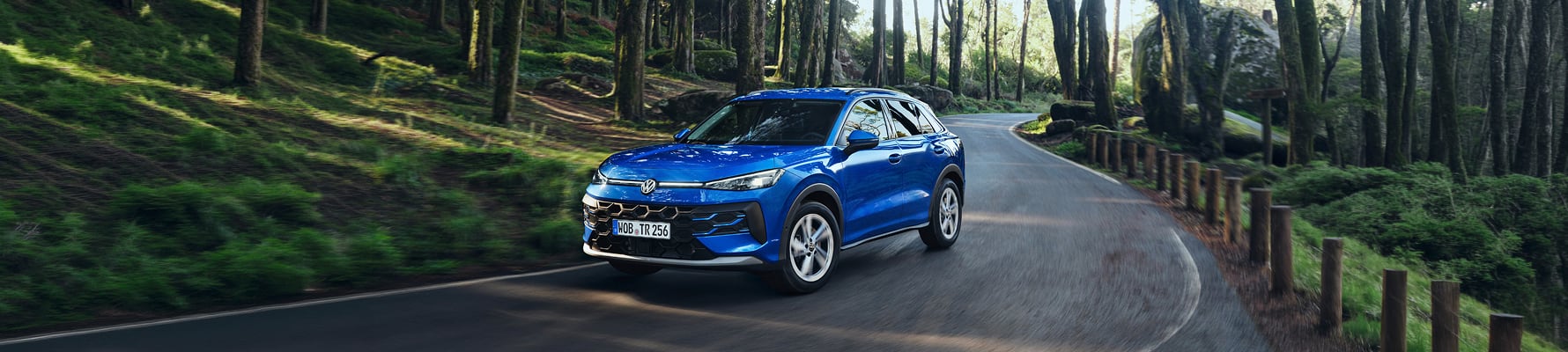 Blue Volkswagen T-Roc driving on a curved forest road with trees and rocks in the background during daytime