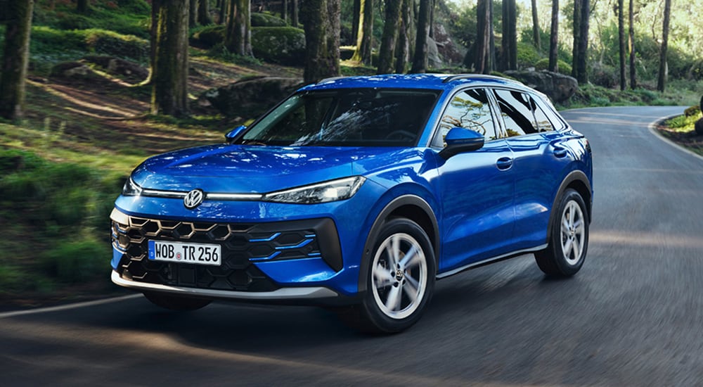 Blue Volkswagen T-Roc driving on a curved forest road with trees and rocks in the background during daytime